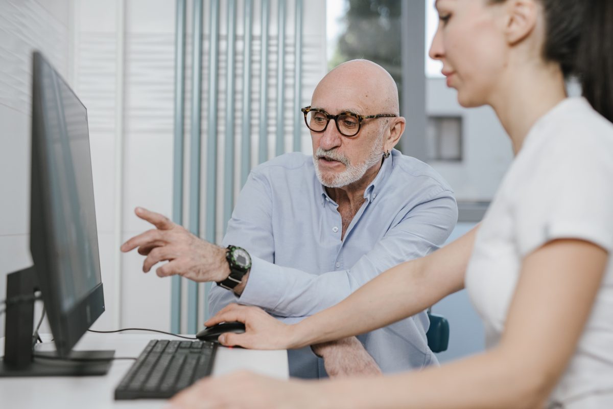 An older man with glasses pointing at a computer screen while a helper shows him what to do